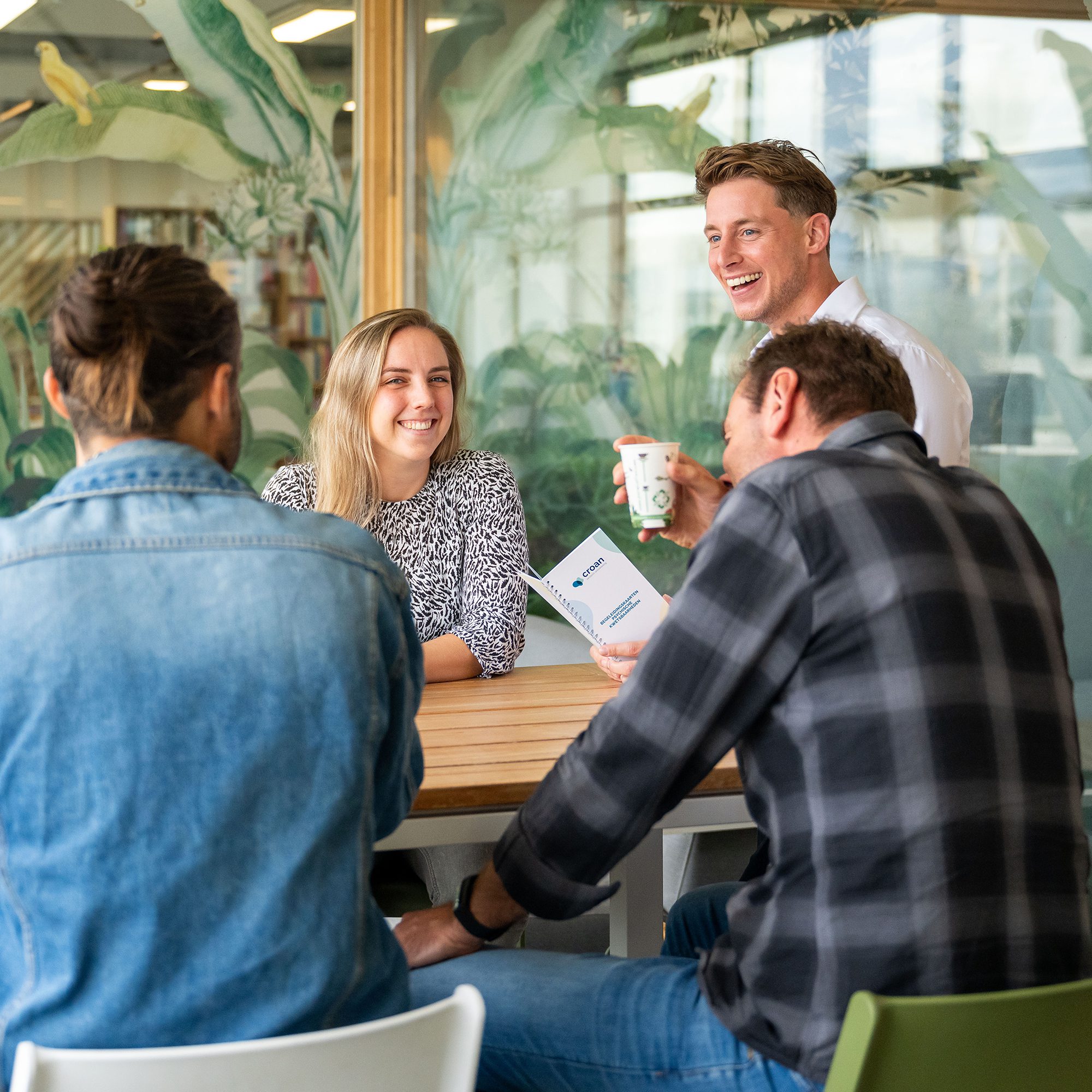 Vier collega's in ontspannen, open gesprek aan een kantoortafel — gedragscode die zichtbaar wordt in de dagelijkse samenwerking